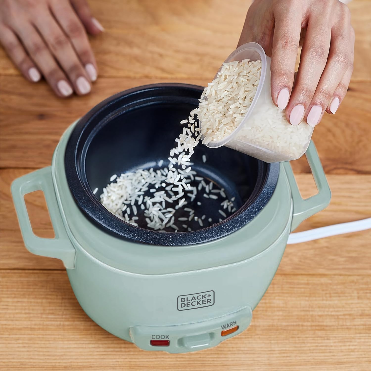 Person pouring rice into a Black+Decker rice cooker on a wooden surface