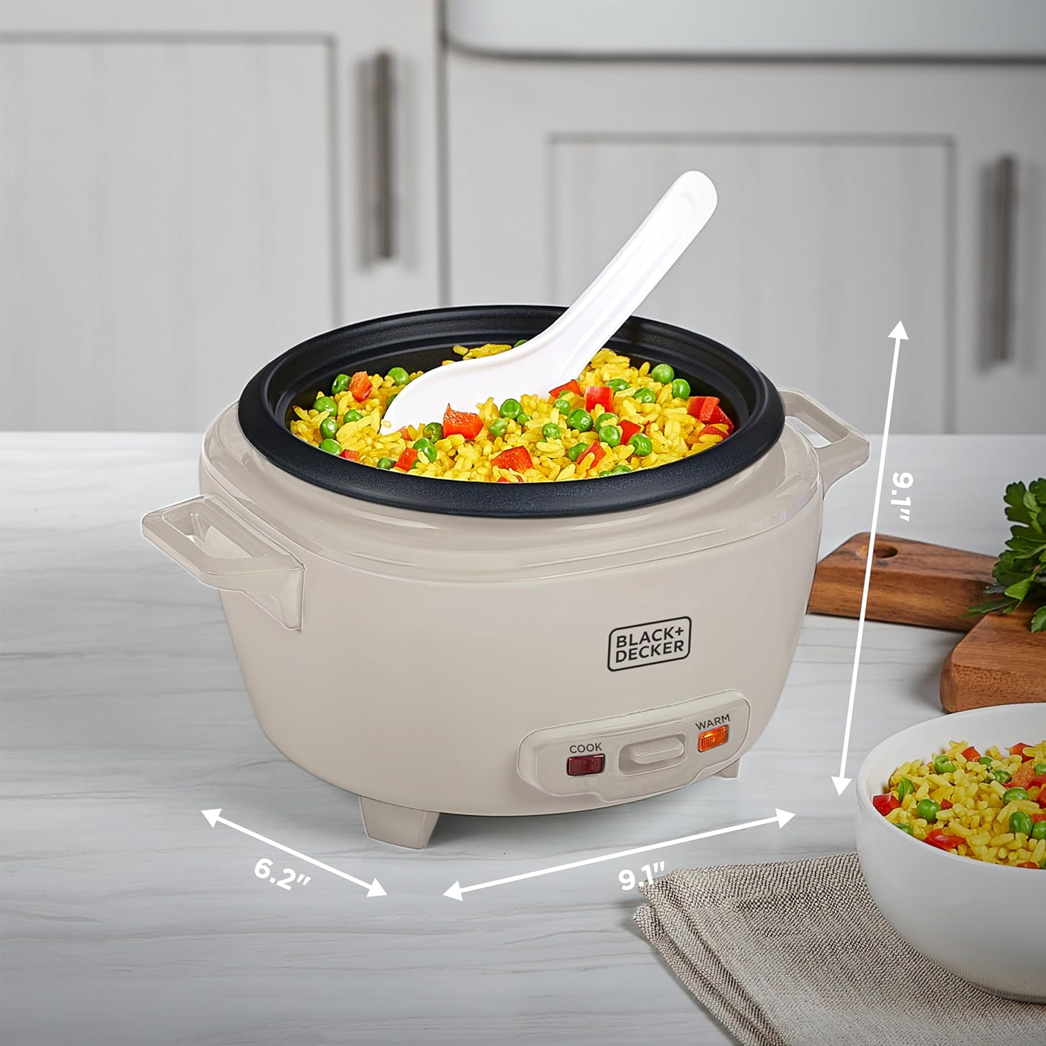 Beige rice cooker with a white spoon inside, filled with colorful rice and vegetables, on a kitchen counter.