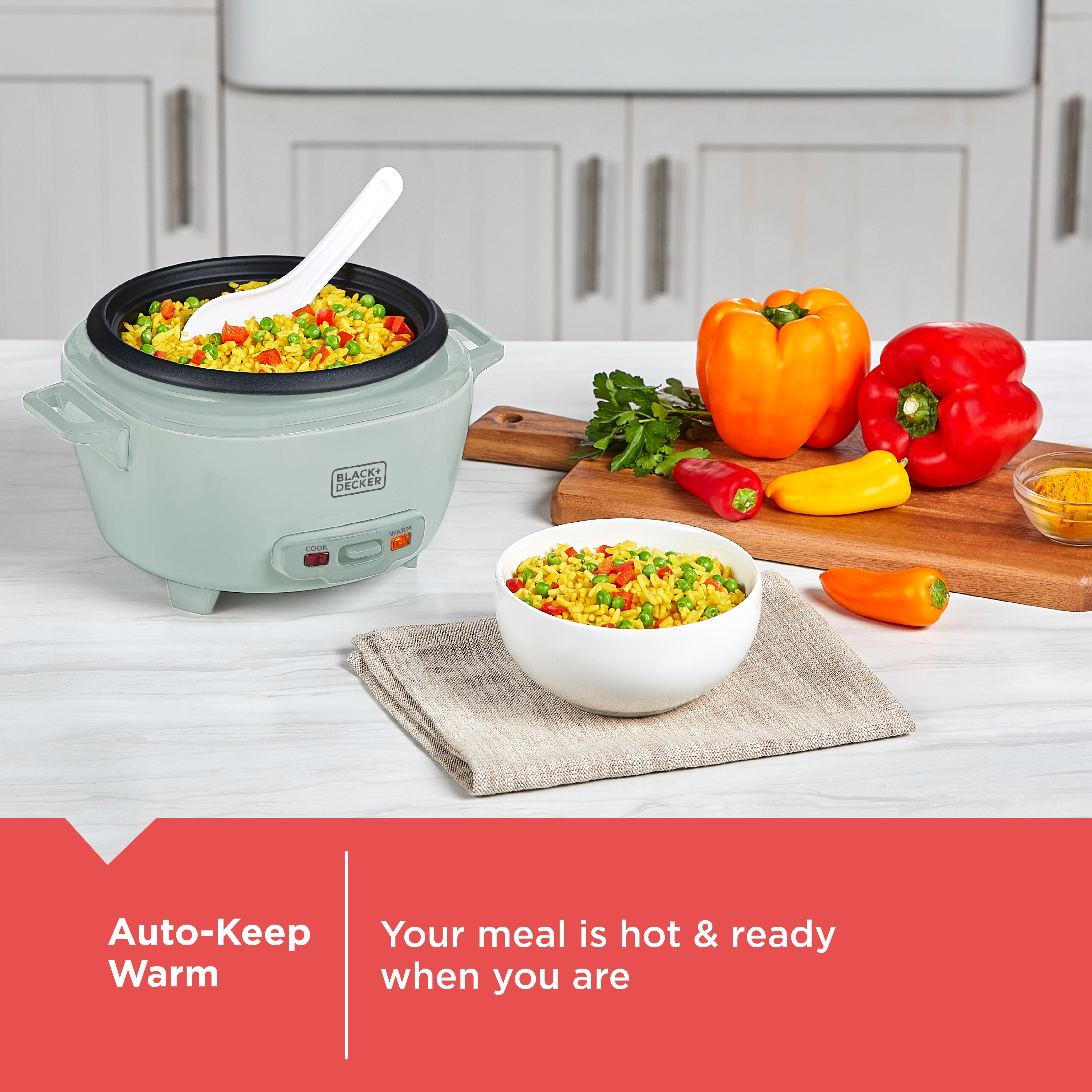 Green rice cooker with a bowl of rice and vegetables on a kitchen counter, surrounded by fresh peppers.