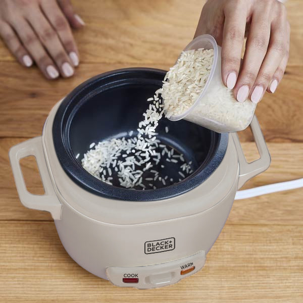 Person pouring rice into a Black+Decker electric rice cooker on a wooden surface