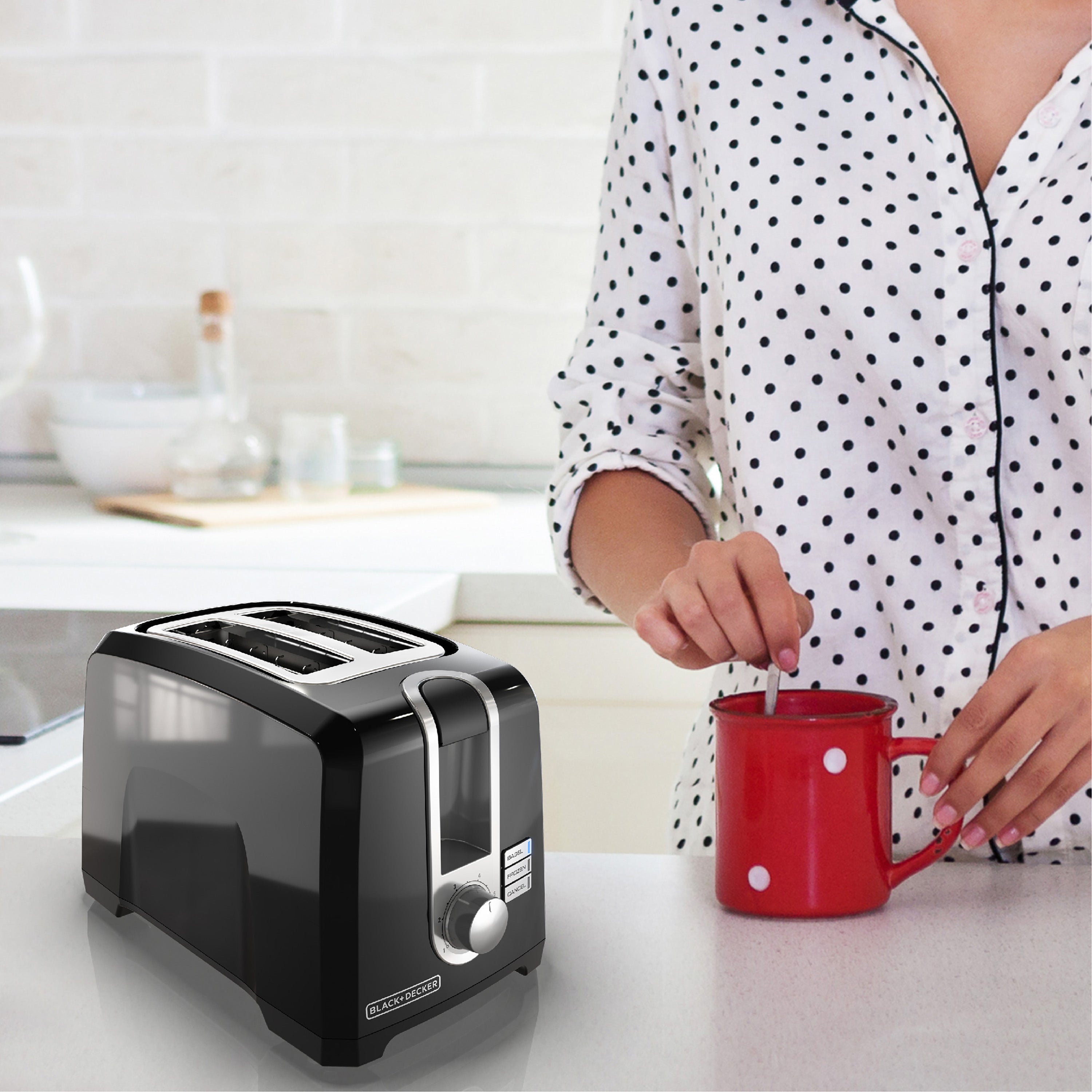 Black toaster on a kitchen counter with a person holding a red mug with white polka dots.