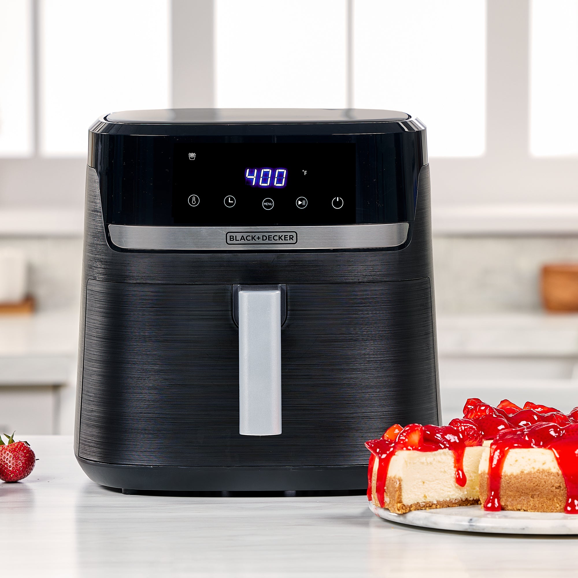 Black air fryer on a kitchen counter with a cheesecake and strawberries in the foreground