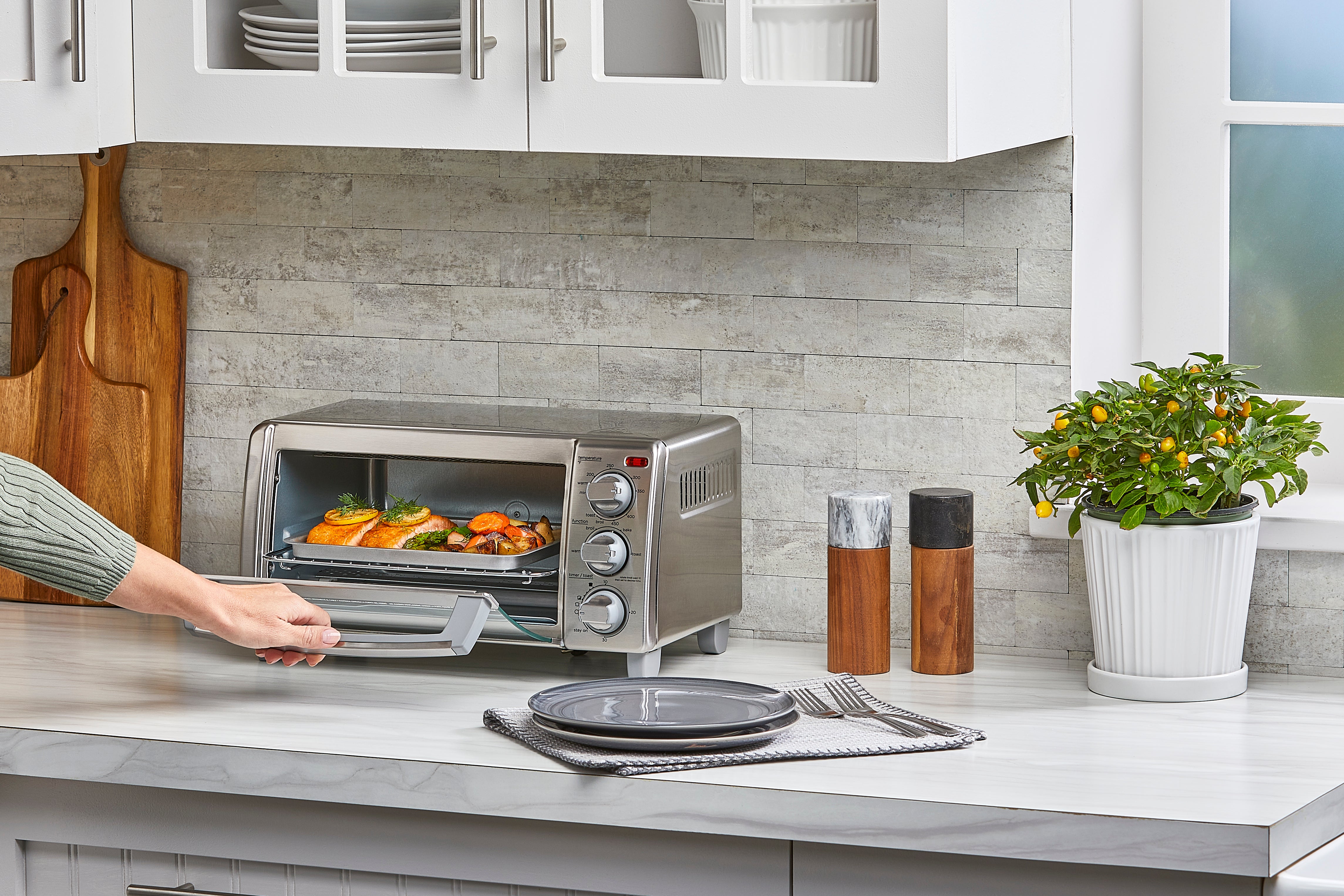 Person using a toaster oven in a modern kitchen with a potted plant and kitchen utensils.