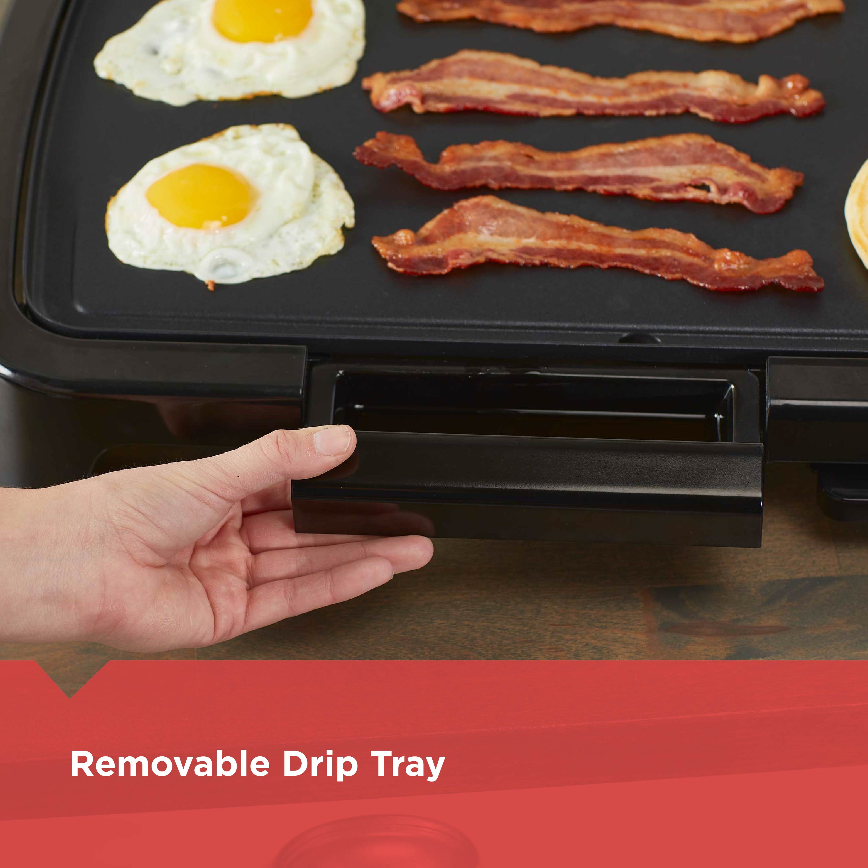 Person holding a black electric grill with eggs and bacon on it, featuring a removable drip tray.
