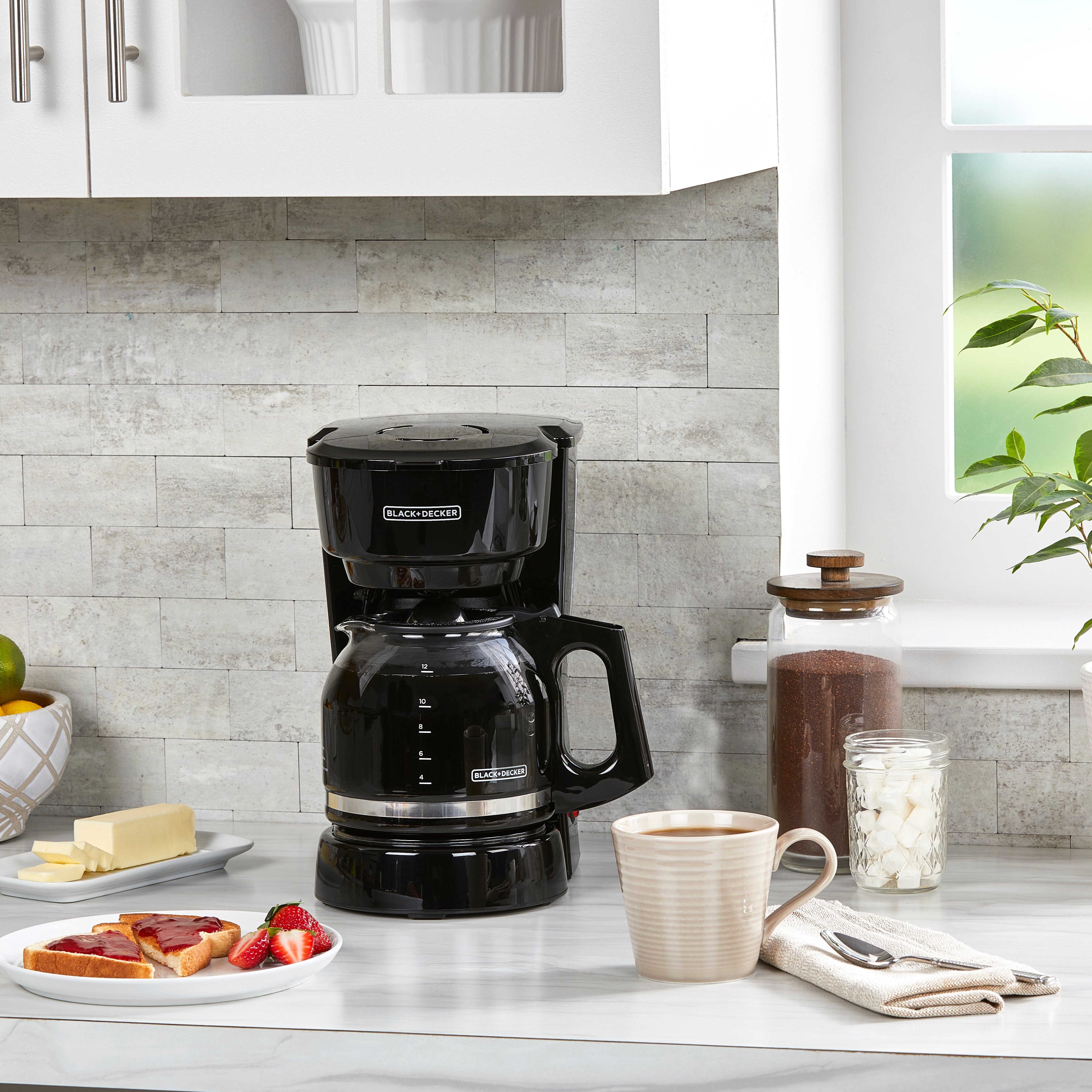 Black coffee maker on a kitchen counter with a cup of coffee, plate of food, and decorative items.