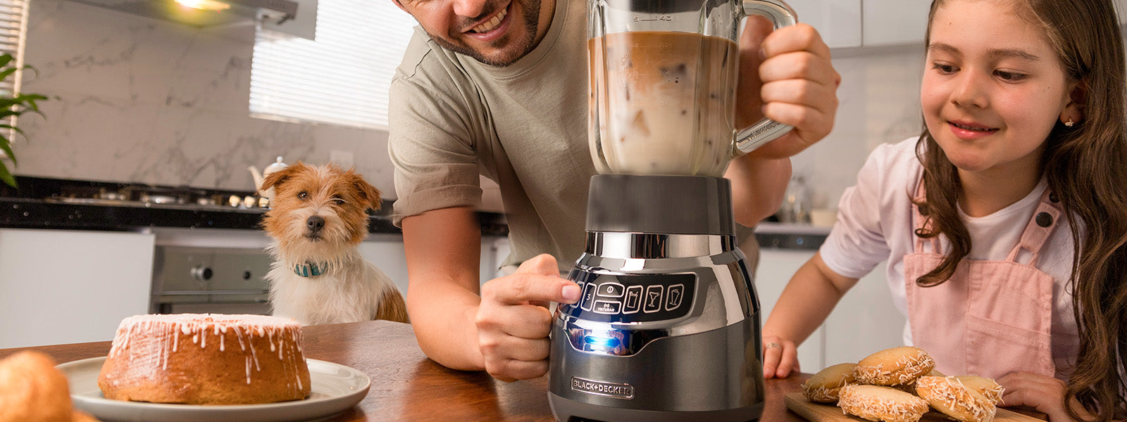 Man and young girl in a kitchen with a blender and dessert, dog in the background