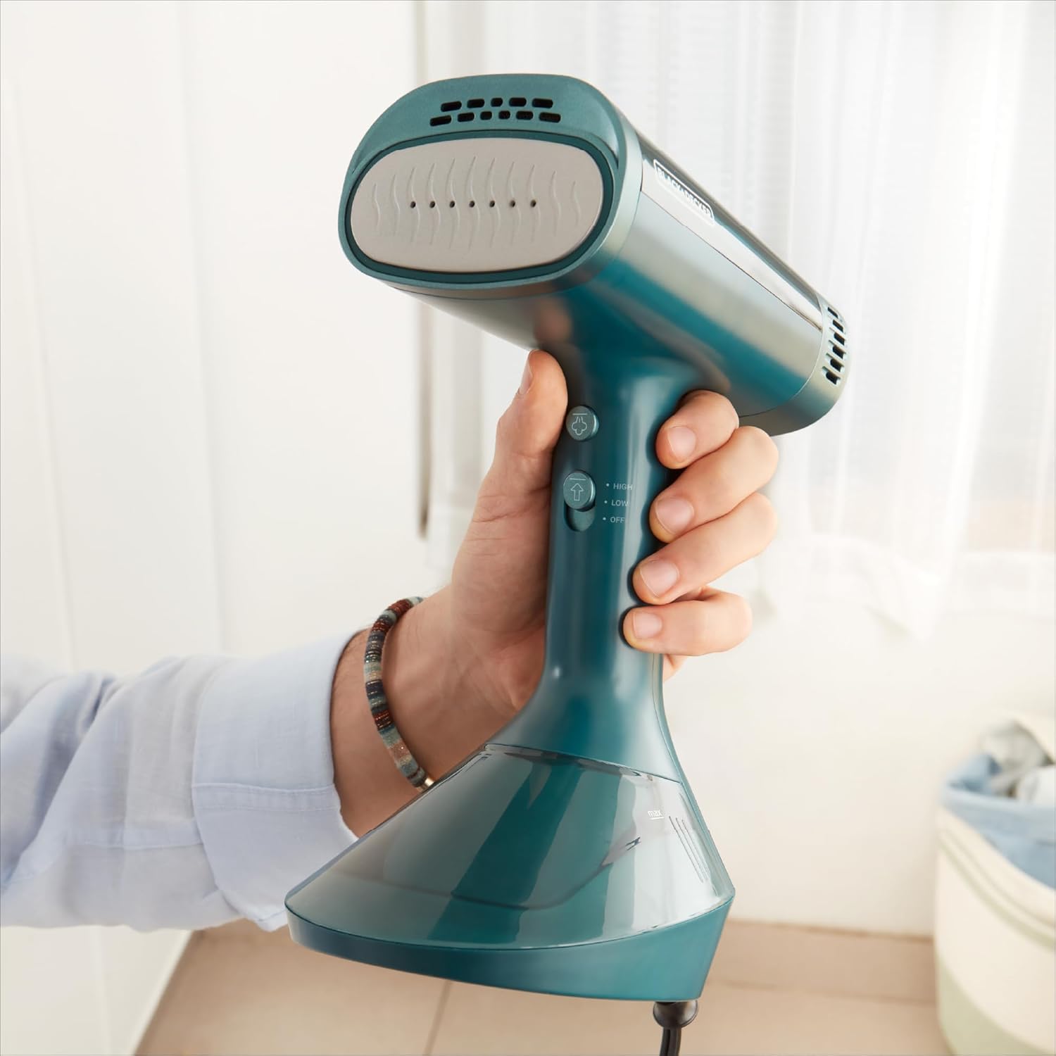 Hand holding a teal handheld steamer against a blurred indoor background