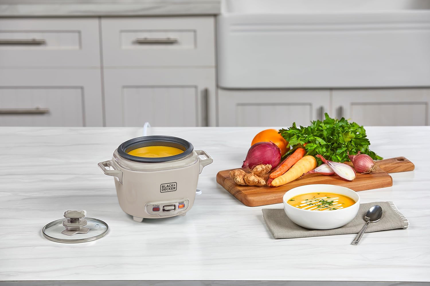 Small rice cooker on a kitchen counter with a bowl of soup and vegetables.