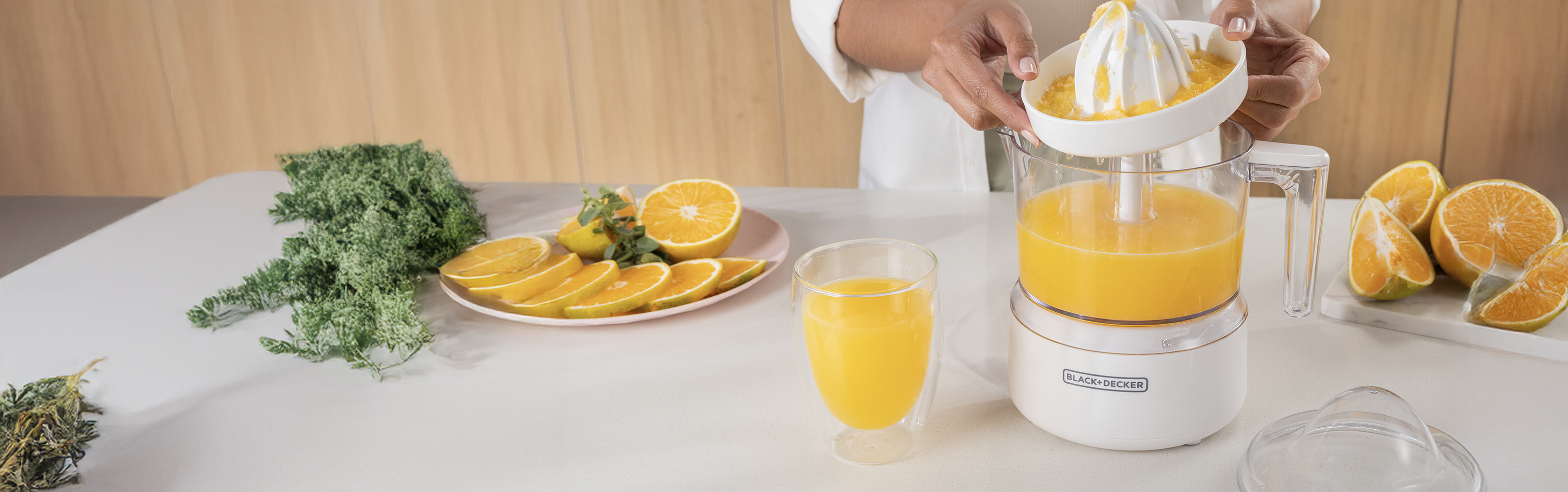 Person using a juicer to extract juice from oranges on a table with a glass of juice and a plate of oranges.