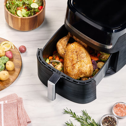 Black air fryer with roasted chicken and vegetables on a wooden table with fresh ingredients.